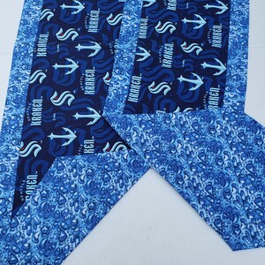 May include: Two blue and white fabric table runners with a repeating pattern of a blue anchor and the words "Seattle Kraken". The fabric has a water drop pattern.
