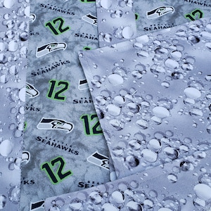 May include: Gray fabric with a repeating pattern of a green and black Seattle Seahawks logo, the number 12, and the words "Seattle Seahawks". The fabric has a white background with a pattern of gray circles.