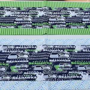 May include: Two table runners with a Seattle Seahawks theme. The top runner has a green and white striped border with a blue background featuring the Seahawks logo and the word "Seahawks" repeated. The bottom runner has a blue background with a white splatter paint effect and the Seahawks logo and the word "Seahawks" repeated.
