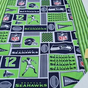 May include: Green and blue fabric with a repeating pattern of Seattle Seahawks logos, footballs, and the number 12. The fabric has a white background and is trimmed with green stitching.