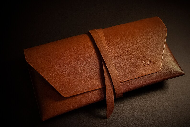 K&ouml;nnte beinhalten: Eine braune Leder-Clutch mit Klappenverschluss und Wickelriemen. Die Clutch hat die Initialen "AA" auf der Vorderseite eingepr&auml;gt. Das Leder hat ein strukturiertes Aussehen.