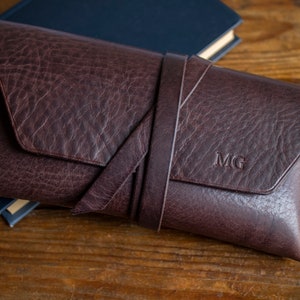 K&ouml;nnte beinhalten: Eine dunkelbraune Leder-Clutch mit strukturierter Oberfl&auml;che und einem umlaufenden Riemen. Die Initialen "MG" sind auf der Vorderseite eingepr&auml;gt. Die Clutch liegt auf einer Holzoberfl&auml;che neben einem geschlossenen blauen Buch.