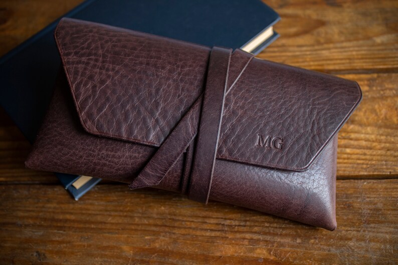 K&ouml;nnte beinhalten: Eine dunkelbraune Leder-Clutch mit strukturierter Oberfl&auml;che und einem umlaufenden Riemen. Die Initialen "MG" sind auf der Vorderseite eingepr&auml;gt. Die Clutch liegt auf einer Holzoberfl&auml;che neben einem geschlossenen blauen Buch.