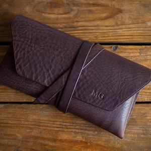 K&ouml;nnte beinhalten: Eine dunkelbraune Leder-Clutch mit einem umlaufenden Riemen, auf einer Holzoberfl&auml;che. Die Initialen "MG" sind eingepr&auml;gt. Das Leder hat eine strukturierte Optik.