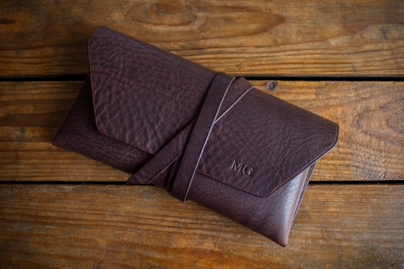 K&ouml;nnte beinhalten: Eine dunkelbraune Leder-Clutch mit einem umlaufenden Riemen, auf einer Holzoberfl&auml;che. Die Initialen "MG" sind eingepr&auml;gt. Das Leder hat eine strukturierte Optik.