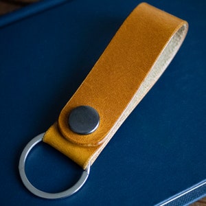 Könnte beinhalten: Ein Schlüsselanhänger aus hellbraunem Leder mit einem silbernen Schlüsselring und einem dunkelgrauen Druckknopf. Der Schlüsselanhänger liegt auf einer dunkelblauen Oberfläche.