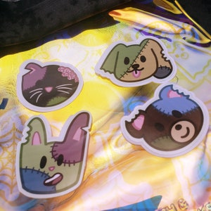 May include: Four cartoon stickers featuring zombie-themed animals: a cat, a dog, a bunny, and a bear. Each sticker has a stitched-together appearance with a mix of colors including green, purple, blue, and brown. The cat has exposed brains, and the dog has its tongue out.