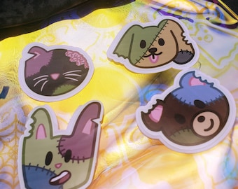 Peut inclure: Quatre autocollants de dessins animés représentant des animaux sur le thème des zombies : un chat, un chien, un lapin et un ours. Chaque autocollant a une apparence cousue avec un mélange de couleurs, dont le vert, le violet, le bleu et le marron. Le chat a le cerveau exposé et le chien tire la langue.