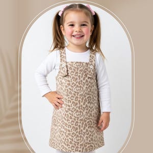Puede incluir: Una niña lleva un vestido con estampado de leopardo marrón y beige sobre una camisa blanca de manga larga. El vestido tiene tirantes abotonados. El texto de la imagen dice "KIDS DRESS SEWING PATTERN" y "4Y-8Y".