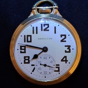 May include: A gold-toned pocket watch with a white face, featuring black numerals and hands. The watch face displays the brand name "HAMILTON". A smaller subdial is visible at the bottom. The watch has a loop at the top for a chain.