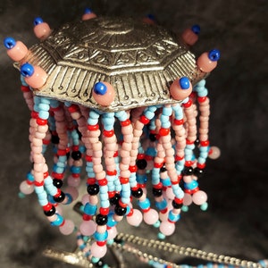 May include: A silver-toned, ornate headpiece with a geometric design. It features dangling strands of pink, blue, red, and black beads. The headpiece has a central, decorative element with a series of pink and blue beads.