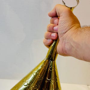 May include: A gold-colored bottle opener with a flared, textured design. The opener has a ring at the top for gripping and a decorative, scale-like pattern on the body. The item is held in a hand.