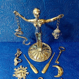 Könnte beinhalten: Eine Sammlung goldfarbener Metallanhänger und -figuren auf blauem Hintergrund. Artikel umfassen eine Statue einer Frau mit Waage, eine Meerjungfrau, eine Sonne mit Gesicht, eine Rettungsboje, einen Anker, ein Steuerrad, einen Schlüssel und einen Mond.