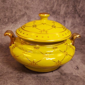 May include: A yellow ceramic lidded serving dish with gold accents. The dish features a repeating gold starburst pattern and gold trim on the handles and lid. The lid has a gold knob.