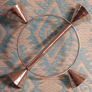 May include: A silver-colored, decorative metal object with a circular frame and a central rod. Four cone-shaped elements with etched designs are attached to the frame and rod, creating a symmetrical design. The object is displayed against a blue and white patterned background.