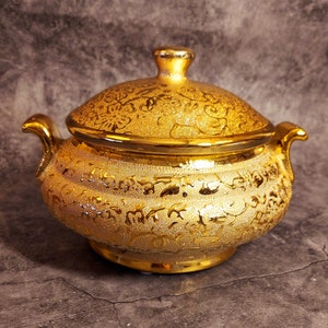 May include: A gold-coloured, lidded serving dish with detailed embossed patterns. The dish features two handles and a knob on the lid. The design is ornate and decorative, suitable for serving food.