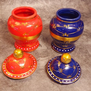 May include: Two decorative ceramic jars, one red and one blue, with matching lids. Each jar features gold bands and starburst designs. The lids have gold knobs and are decorated with gold and white accents. The jars are approximately 4 inches tall.