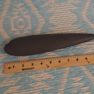 May include: A dark gray, elongated object with a rounded end and a pointed tip. A wooden ruler with inch markings is placed next to the object. The ruler reads "WESTCOTT".