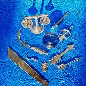 May include: An assortment of silver-toned miniature charms and pendants, including a balance scale, swords, arrows, a feather, and a machete, arranged on a textured blue surface. The collection evokes themes of justice, weaponry, and fantasy.