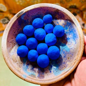 May include: A ceramic bowl with a speckled, light brown exterior and a blue and purple interior holds a collection of vibrant blue spheres. The spheres are evenly spaced and fill the bowl, creating a striking contrast of colors and textures.