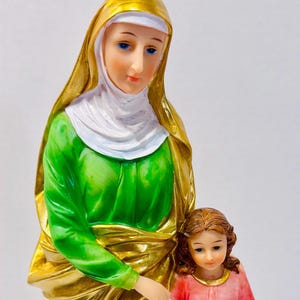 May include: A religious figurine depicting a woman in a gold head covering and green garment, with a young girl in a pink dress. The woman has blue eyes and is holding the girl's hand. The girl is holding a book.