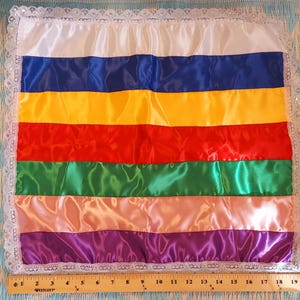 Puede incluir: Una almohada rectangular de satén con rayas horizontales en colores del arcoíris: blanco, azul, amarillo, rojo, verde, melocotón y morado. La almohada está bordeada con encaje blanco y mide unos 30 cm de ancho.
