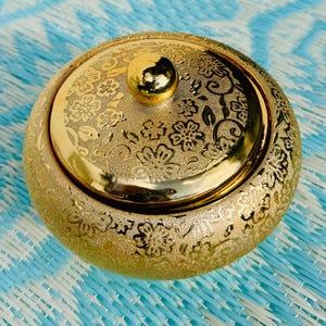 Pode incluir: Um recipiente redondo de cor dourada com uma tampa a condizer que apresenta um intrincado padrão floral em relevo. A tampa é rematada com um botão esférico dourado liso e polido. Toda a superfície do recipiente tem um acabamento texturizado.