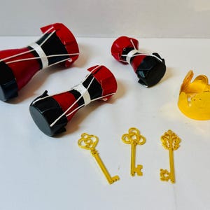 May include: A collection of miniature decorative items, including three red, black, and white drums, three gold-colored keys, and a gold-colored crown-shaped object. The drums have a cylindrical shape.