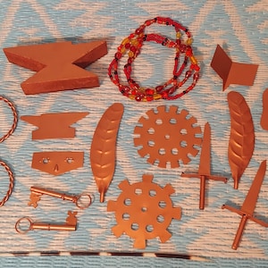 Puede incluir: Un surtido de suministros de artesanía de metal color cobre, que incluyen un yunque, llaves, plumas, engranajes, espadas y un collar de cuentas. Los artículos están dispuestos sobre una superficie estampada azul y blanca.