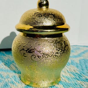 May include: A gold-colored ceramic jar with a matching lid, featuring a textured surface with embossed floral and vine patterns. The jar has a rounded body and a smooth, reflective finish on the lid knob and rim, creating a metallic appearance.