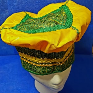 Può includere: Un cappello di raso giallo con finiture verdi e dorate. Il cappello ha una corona arrotondata e una fascia decorativa alla base. Le finiture includono pizzo e dettagli dorati.
