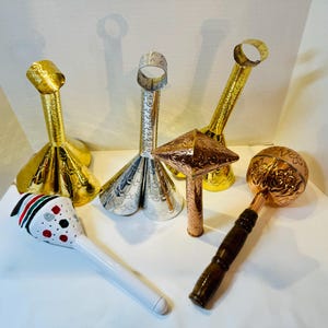 May include: A collection of six musical instruments including three metallic cone-shaped bells with ring handles, one white maraca with red, green, and black dots, one copper-coloured star-shaped percussion instrument, and one copper-coloured round rattle.