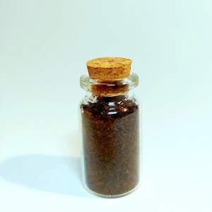 May include: A small, clear glass bottle filled with dark brown material, sealed with a cork stopper. The bottle's contents appear to be finely shredded. The background is a plain white surface.