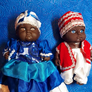 May include: Two dolls with dark skin and detailed facial features. One doll wears a blue and white outfit with a matching head wrap. The other doll is dressed in a red jacket, white pants, and a red and white head covering. Both dolls have detailed clothing.
