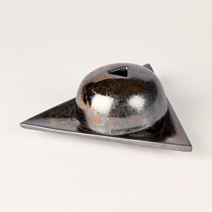 May include: A ceramic sculpture featuring a dark, metallic-glazed sphere with a triangular opening, resting on a triangular base. The sphere has a textured surface with hints of bronze and white. The base is a dark, matte finish.