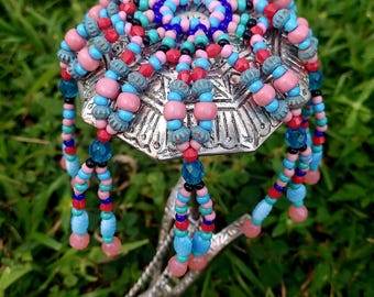 Może przedstawiać: Srebrna łyżka do serwowania z wzorem z koralików. Miseczka łyżki ozdobiona jest kolorową gamą różowych, niebieskich, czerwonych i czarnych koralików. Wiszące pasma koralików dodają uroku dekoracyjnego. Łyżka jest ustawiona na tle zielonej trawy.