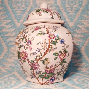 May include: A white ceramic jar with a lid, adorned with a floral pattern in pink, blue, yellow, and green. The jar features a detailed design of flowering branches and is likely used for decorative purposes.