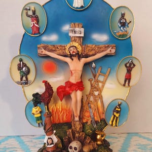 Peut inclure: Figurine religieuse représentant la crucifixion de Jésus, entourée de sept figures dans des cadres circulaires. La base porte l'inscription "7 Potencias Africanas". La palette de couleurs comprend l'or, le rouge, le bleu et le marron.