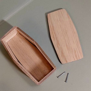 Könnte beinhalten: Eine offene, unfertige Holzkiste mit separatem Deckel und zwei kleinen Nägeln. Die Kiste ist rechteckig mit abgerundeten Enden, und der Deckel hat die gleiche Form. Das Holz hat eine hellbraune Farbe.