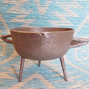 May include: A small, cast iron bowl with three legs and two handles. The bowl is a dark grey colour and has a rough texture. The handles are triangular in shape. The bowl is sitting on a blue and white woven surface.