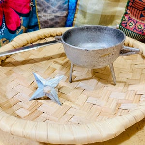 May include: A small, silver-coloured metal cooking pot with three legs and two handles sits on a woven bamboo tray. A silver-coloured metal knife and a star-shaped object are also on the tray. The tray has a woven pattern and a rounded edge.