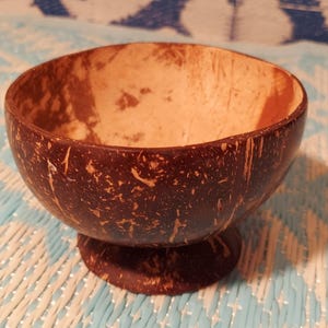 May include: A handcrafted coconut bowl with a pedestal base. The bowl's exterior is a dark brown, textured surface, while the interior is a lighter tan color. This natural bowl is suitable for serving food or as a decorative item.