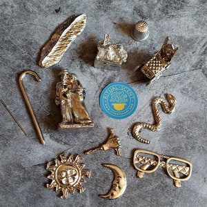 Op de afbeelding: Een assortiment kleine, goudkleurige metalen bedels en objecten, waaronder een veer, een huis, een vingerhoed, een wandelstok, een figuur van twee personen, een slang, een hand die een gereedschap vasthoudt, een zon, een halve maan en een bril. Er is ook een blauwe sticker met witte tekst.