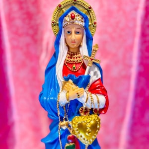 May include: A religious figurine of a woman in a blue and red robe, with gold jewellery and a crown. She holds a golden cross and a heart-shaped pendant. The figure has a halo and is set against a pink backdrop.