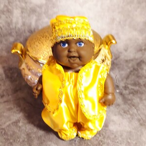 May include: A doll with dark skin, blue eyes, and a smiling expression. The doll is dressed in a bright yellow satin outfit with gold trim, including a hat and vest. The doll is positioned in front of a gold-colored decorative object.