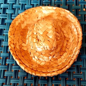 Puede incluir: Un pequeño sombrero de paja tejido con una corona redondeada y un ala ancha ligeramente curvada. El sombrero es de color marrón claro y parece estar hecho de fibras naturales. El sombrero está sobre una superficie tejida azul y turquesa.
