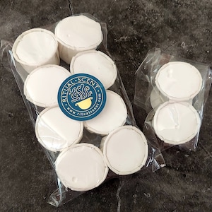 May include: Several white, round wax melts in clear plastic packaging. A blue and gold sticker with the text "RITUAL SCENT" is visible. The melts are arranged on a dark gray surface.