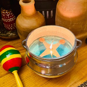 May include: A lit candle in a metallic silver pot with two handles, featuring a star-shaped wick holder and blue wax. Beside it sits a maraca with red, yellow, and green stripes and a yellow handle, near a tan ceramic vase and a large glass jar.