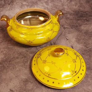 May include: A yellow ceramic serving dish with a lid, featuring gold accents. The dish has two handles and a gold interior. The lid is decorated with a gold starburst pattern and a gold knob. The dish is likely for serving food.