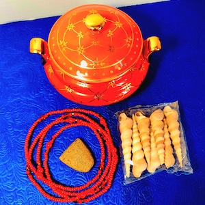 May include: A red ceramic casserole dish with gold accents and a lid, a red beaded necklace with a stone, and a package of seashells on a blue surface. The casserole dish has gold handles.